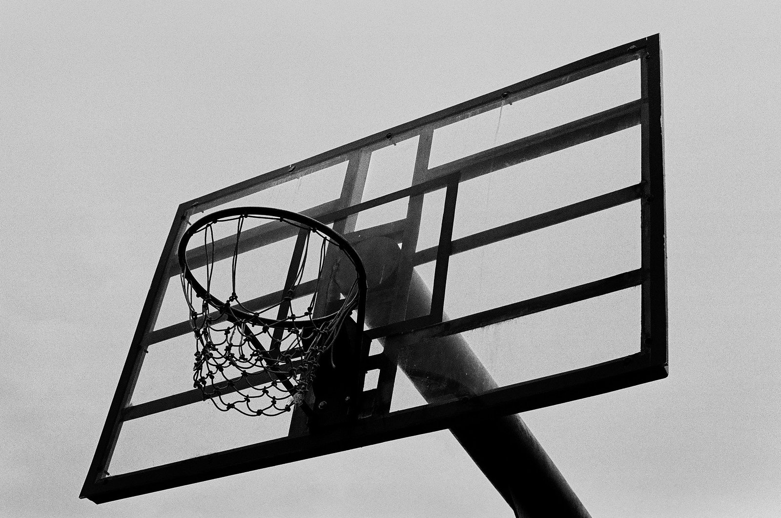 Low angle black and white photo of a basketball hoop and backboard against a gray sky