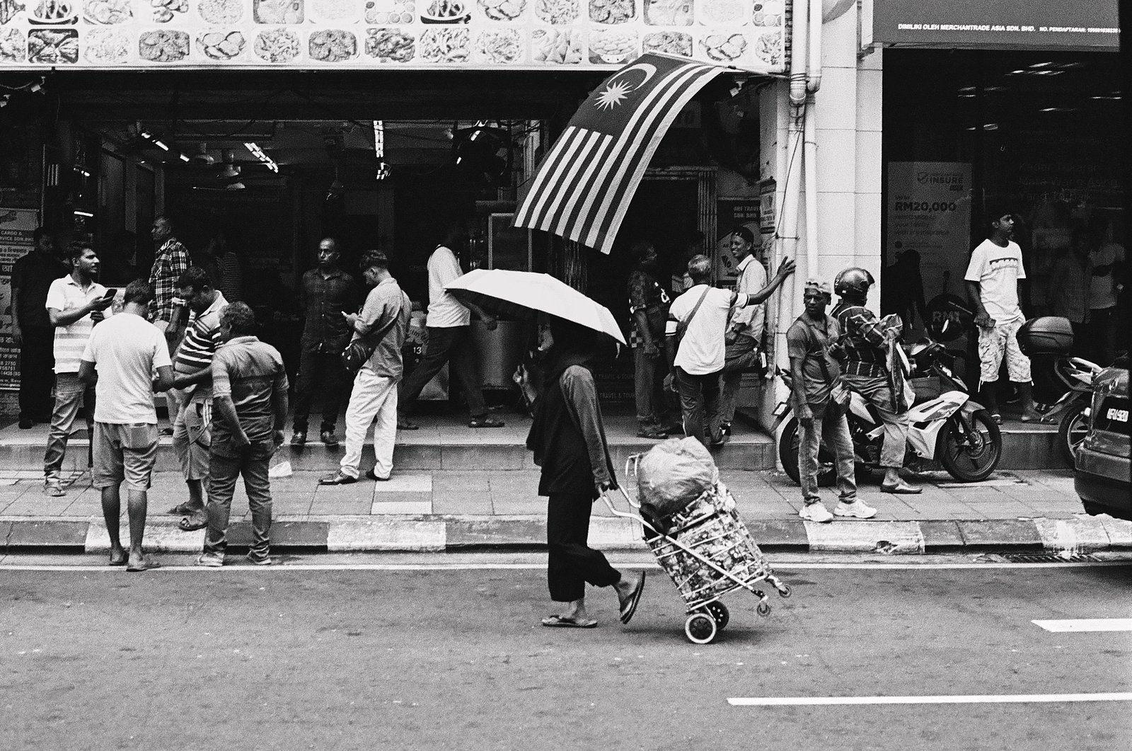Woman pulling a cart under an umbrella on a street in Kuala Lumpur, Malaysia, with a large flag overhead